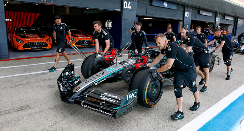 Varios miembros del equipo empujan un carro de Fórmula 1 Mercedes-AMG PETRONAS fuera del pit lane, con dos carros naranjas Mercedes-AMG GT al fondo.