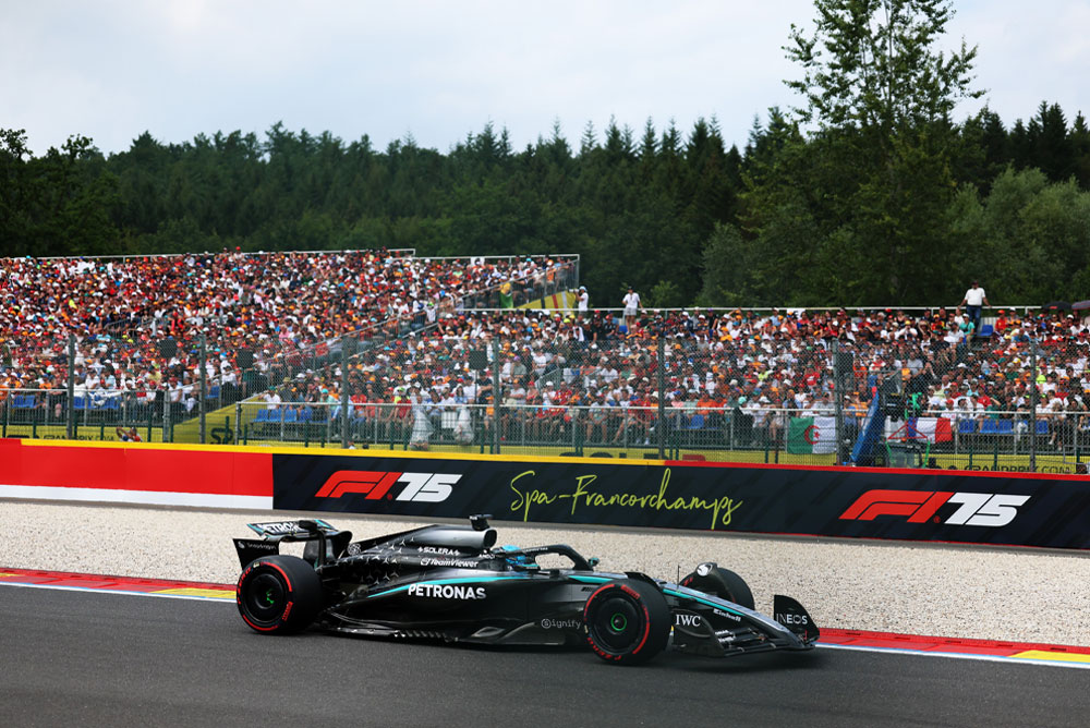 Un auto del equipo Mercedes-AMG PETRONAS Formula One Team corre en el circuito de Spa-Francorchamps. Al fondo, las tribunas llenas están repletas de aficionados que agitan banderas de diferentes países. El logo de Spa-Francorchamps se puede ver en la barrera.