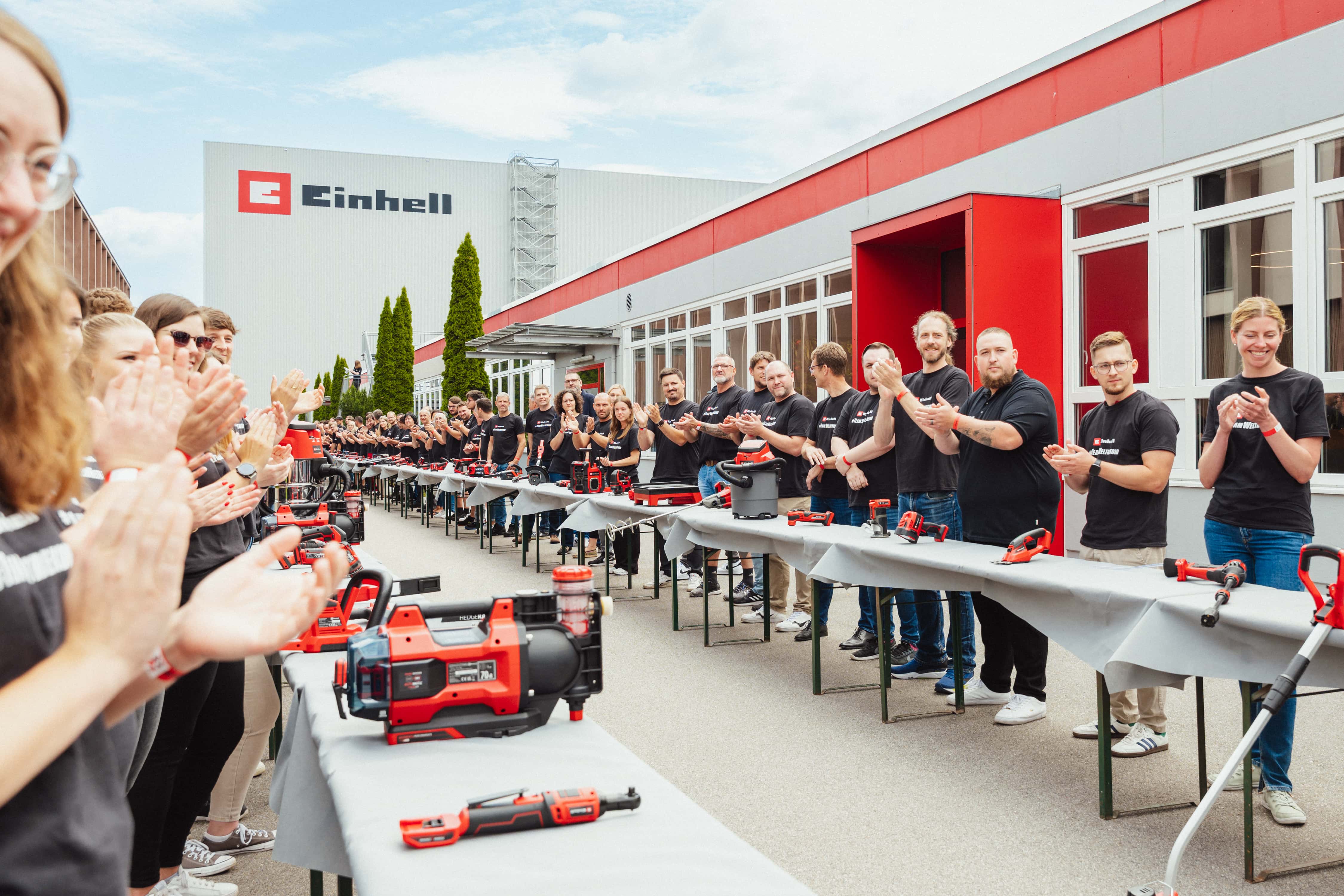 Numerosos empleados de Einhell aplauden junto a una larga fila de mesas con herramientas inalámbricas en las instalaciones de la fábrica.